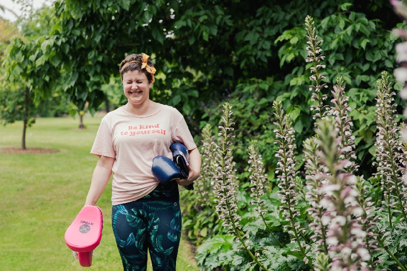 A female personal trainer is holding boxing gloves and squinting into the sun in a garden